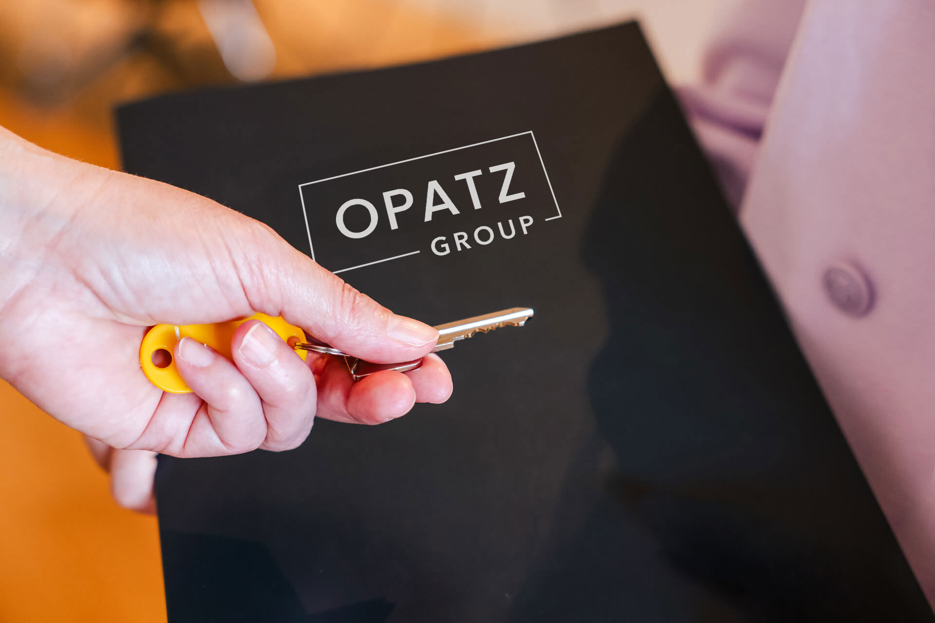 Eine Hand übergibt einen Schlüssel, im Hintergrund ist eine Mappe mit dem Logo der OPATZ Group zu sehen.
